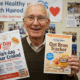 Prompt ảnh: Friendly elderly man (Harold) smiling while holding a printed “30-Day Oat Bran Tracker”. Behind him, wall with pinned photos of group members, heart icons, breakfast photos. Banner text: “Live Healthy with Harold – Join the Journey”. Inviting, authentic tone.